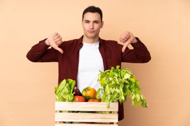 Bej arka planda izole edilmiş bir kutuda taze sebze toplayan çiftçi baş parmağını aşağı gösteriyor.