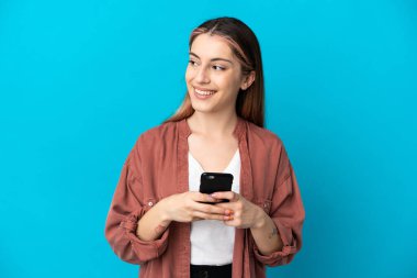 Kafkasyalı genç bir kadın mavi arka planda izole edilmiş ve cep telefonu kullanıyor.