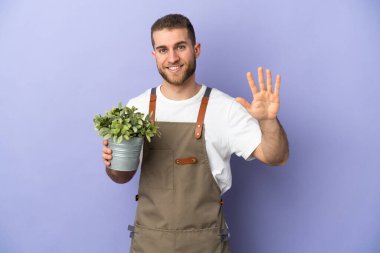 Bahçıvan Kafkasyalı adam sarı arka planda bir bitki tutuyor, parmaklarıyla beşi sayıyor.