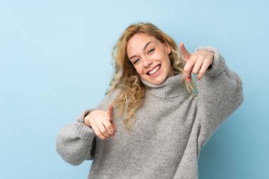 Mavi arka planda izole edilmiş süveter giyen genç sarışın kadın gülümserken seni işaret ediyor.