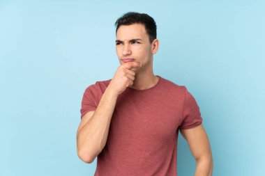 Kafkasyalı yakışıklı bir adam mavi arka planda izole edilmiş. Şüpheleri var ve kafası karışmış bir yüz ifadesi var.