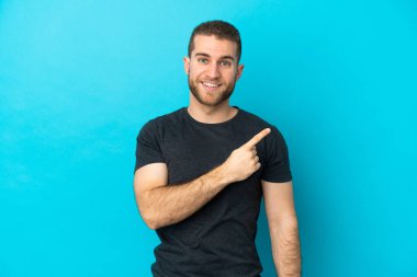 Mavi arka planda izole edilmiş yakışıklı beyaz adam bir ürünü sunmak için kenarı işaret ediyor.