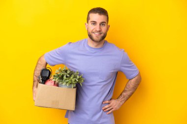 Beyaz bir genç, sarı arka planda izole edilmiş eşyalarla dolu bir kutuyu kaldırırken kalçasında kollarıyla poz veriyor ve gülümsüyor.