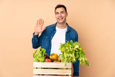 Taze toplanmış sebzeli çiftçi bej arka planda izole edilmiş bir kutuda mutlu bir ifadeyle selamlıyor.