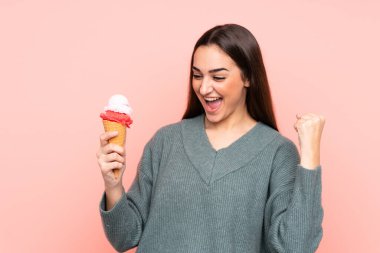 Kornet dondurması tutan genç bir kadın pembe arka planda izole edilmiş bir şekilde zaferi kutluyor.