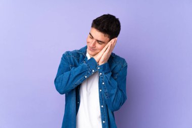 Mor arka planda izole edilmiş genç, beyaz, yakışıklı bir adam uyku jesti yapıyor.