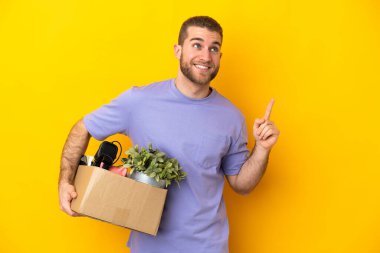 Genç Kafkasyalı, sarı arka planda izole edilmiş eşyalarla dolu bir kutuyu seçerken harika bir fikre işaret ediyor.