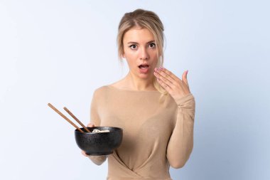 Sarışın kadın, izole edilmiş mavi arka planda elinde çubuklu erişte kasesiyle şaşırmış.