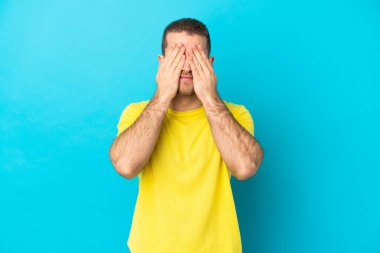Genç, yakışıklı, beyaz bir adam mavi arka planda izole edilmiş. Gözlerini elleriyle kapatıyor.