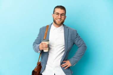 Kafkasyalı genç işadamı mavi arka planda izole edilmiş, kolları kalçasında ve gülümsüyor.