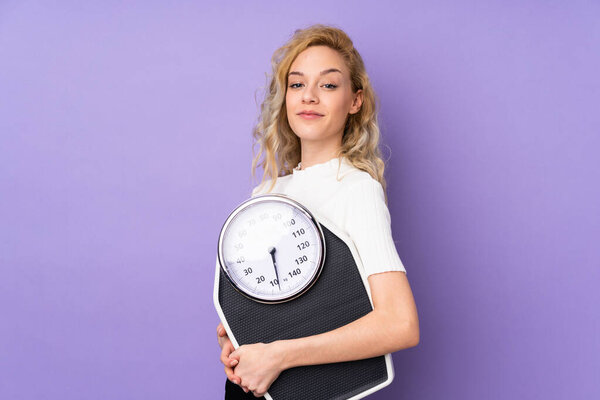 Young blonde woman isolated on purple background with weighing machine