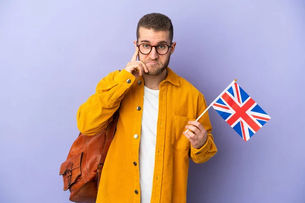 年轻的高加索人拿着一面英国国旗 与紫色背景隔离 思考着一个想法 — 图库照片