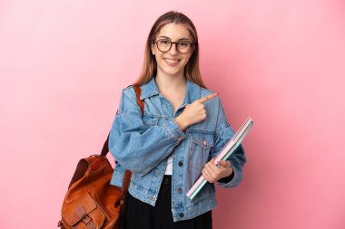 Pembe arka planda izole edilmiş genç beyaz kadın bir ürünü sunmak için kenarı işaret ediyor.