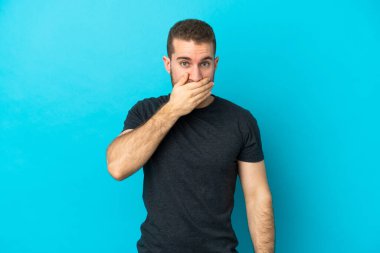Genç, yakışıklı, beyaz bir adam mavi arka planda izole edilmiş, ağzı elle kapatılmış.