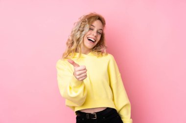 Pembe arka planda izole edilmiş eşofman üstü giyen genç sarışın kadın. Başparmakları havada çünkü iyi bir şey oldu.