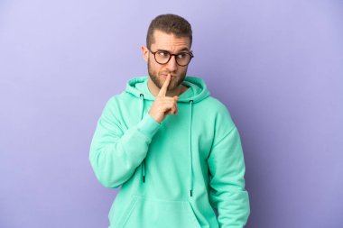 Mor arka planda izole edilmiş yakışıklı beyaz adam sessiz bir jest işareti gösteriyor. Parmağını ağzına sokuyor.
