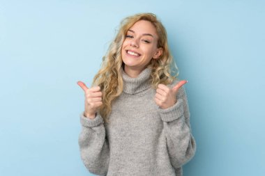 Mavi arka planda izole edilmiş süveter giyen genç sarışın bir kadın. Baş parmakları havada ve gülümsüyor.