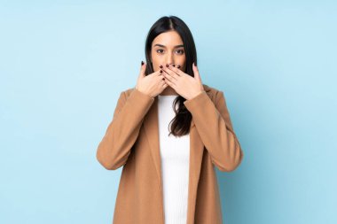 Mavi arka planda izole edilmiş genç beyaz kadın elleriyle ağzını kapatıyor.
