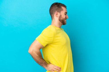 Genç, beyaz, mavi arka planda izole edilmiş bir adam gayret gösterdiği için sırt ağrısı çekiyor.