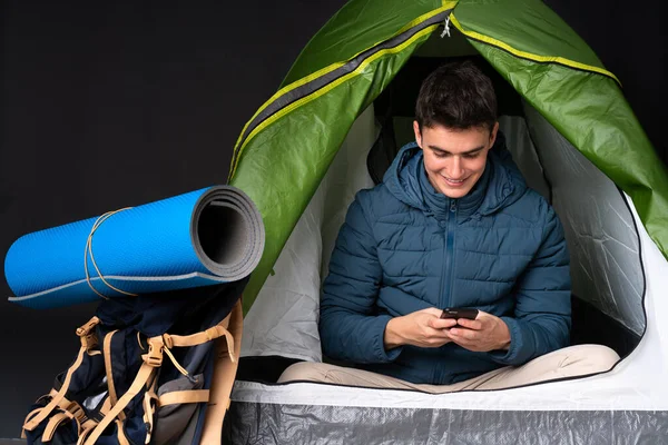 Teenager caucasian man inside a camping green tent isolated on black background sending a message with the mobile