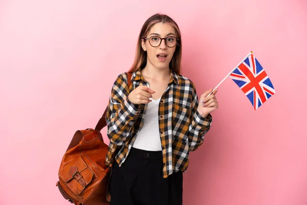 年轻的惊慌失措的女人拿着一面英国国旗 粉色背景孤立 惊讶地指着前方 — 图库照片