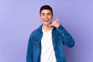 Mor arka planda izole edilmiş genç, beyaz bir adam telefon jesti yapıyor. Bana geri dönüş işareti ver.