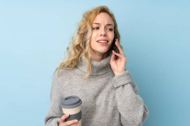 Mavi arka planda izole edilmiş süveter giyen genç sarışın bir kadın. Elinde kahve ve cep telefonu var.