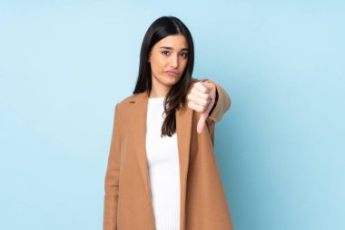 Mavi arka planda izole edilmiş genç beyaz kadın olumsuz ifadeyle baş parmağını aşağı gösteriyor.