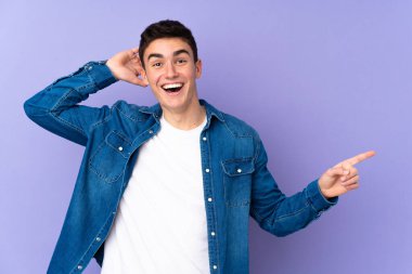 Mor arka planda izole edilmiş, genç, beyaz, yakışıklı bir adam şaşırmış ve parmağıyla kenarı işaret ediyor.