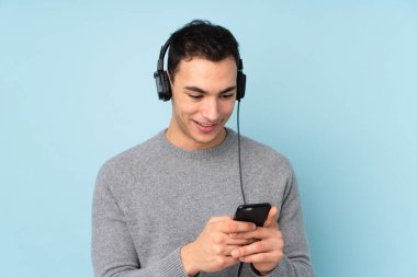 Beyaz tenli yakışıklı adam mavi arka planda izole edilmiş müzik dinliyor ve cep telefonu arıyor.