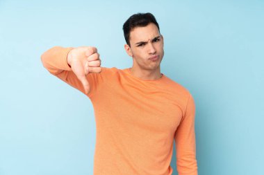 Mavi arka planda soyutlanmış beyaz, yakışıklı genç bir adam, olumsuz bir ifadeyle baş parmağını aşağı gösteriyor.