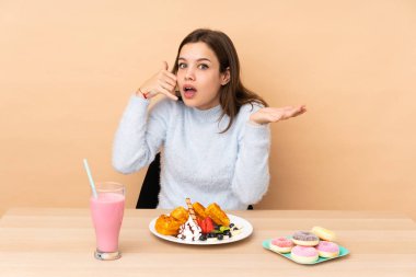 Bej arka planda izole edilmiş waffle yiyen genç bir kız telefon jesti yapıyor ve kuşkulanıyor.