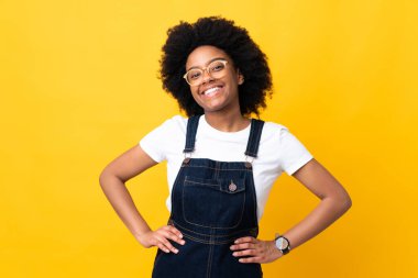 Sarı arka planda izole edilmiş genç Afrikalı Amerikalı kadın kolları kalçasında ve gülümsüyor.