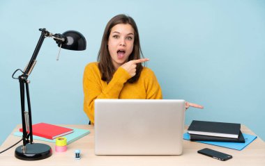 Öğrenci kız evinde çalışıyor. Mavi arka planda izole edilmiş. Şaşırmış ve işaret ediyor.