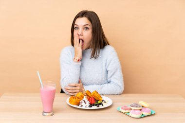 Bej arka planda izole edilmiş waffle yiyen genç kız şaşırmış ve şok olmuş bir yüz ifadesi var.