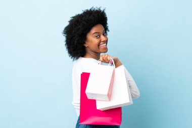 Genç Afrikalı Amerikalı bir kadın mavi arka planda izole edilmiş alışveriş torbaları tutuyor ve gülümsüyor.