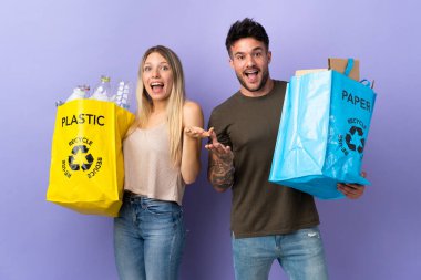 Mor arka planda izole edilmiş, şaşırmış ve şok olmuş bir yüz ifadesiyle genç beyaz çift geri dönüşüm yapıyor.