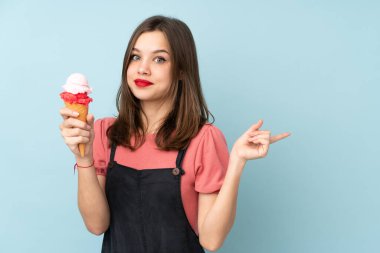 Kornet dondurması tutan genç kız mavi arka planda izole edilmiş, şaşırmış ve parmağıyla işaret ediyor.