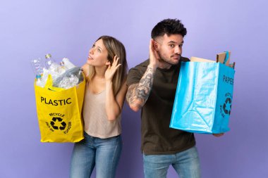 Kafkasyalı genç çift, mor arka planda izole edilmiş bir şekilde geri dönüşüm yapıyor. Kulağına elini sürerek bir şeyler dinliyorlar.