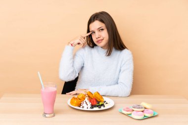 Bej arka planda izole edilmiş waffle yiyen genç kız deliliğin jestini yapıyor. Parmağını kafaya koyuyor.
