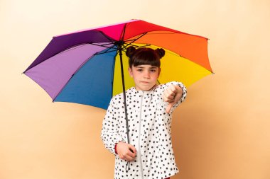 Elinde bej arka planda izole edilmiş bir şemsiye tutan küçük kız, olumsuz ifadelerle baş parmağını aşağı gösteriyor.