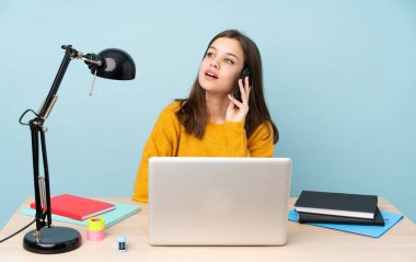 Öğrenci kız, mavi arka planda izole bir şekilde evinde çalışıyor cep telefonuyla sohbet ediyor.
