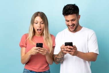 Kafkasyalı genç çift mavi arka planda izole edildi. Şaşırdılar ve cep telefonuyla bir mesaj veya e-posta gönderiyorlar.