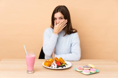 Bej arka planda izole edilmiş waffle yiyen genç kız ağızlarını elleriyle kapatıyor.