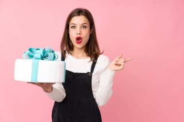 Pembe arka planda büyük bir pasta tutan kız şaşırmış ve işaret ediyor.