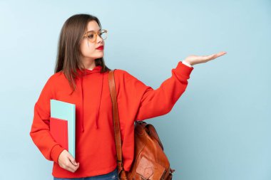 Öğrenci kız mavi arka planda izole kitapları tutuyor. Davet edilmek için ellerini yana açıyor.