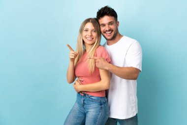 Mavi arka planda izole edilmiş genç beyaz bir çift işaret ediyor ve bir ürün sunuyorlar.