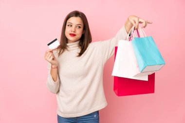 Pembe arka planda izole edilmiş genç bir kız elinde alışveriş çantaları ve bir kredi kartı tutuyor.