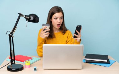 Öğrenci kız, mavi arka planda izole bir şekilde evinde ders çalışıyor. Elinde kahve ve cep telefonu var.