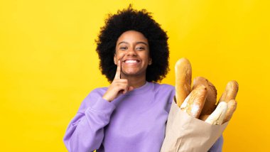 Genç Afrikalı Amerikalı bir kadın sarı arka planda izole edilmiş bir şey alıyor mutlu ve hoş bir ifadeyle gülümsüyor.